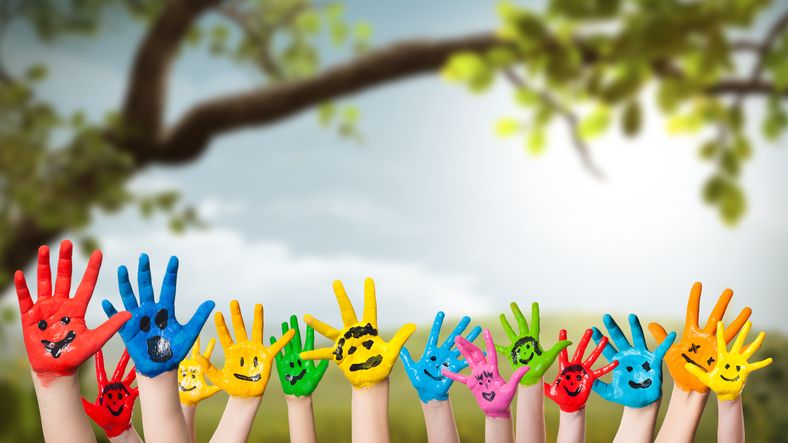 Bund bemalte Kinderhände mit schwarzen lachenden Smileys auf den bunten Händen, die in die Luft gestreckt werden; vor einer Wiese und dem Ast eines Baumes
